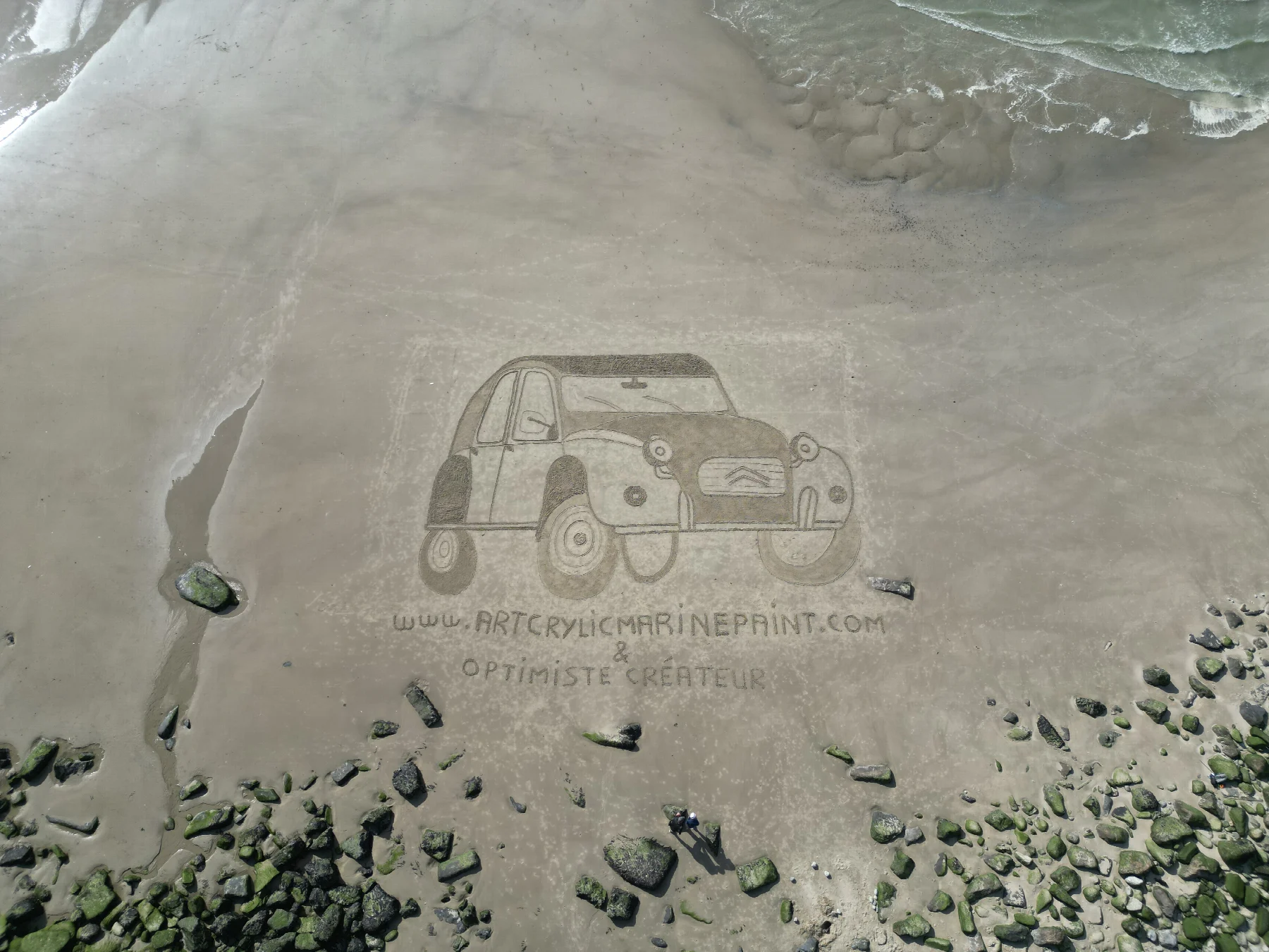 Vue aérienne d’une fresque représentant une voiture ancienne tracée dans le sable entre les rochers et la mer
