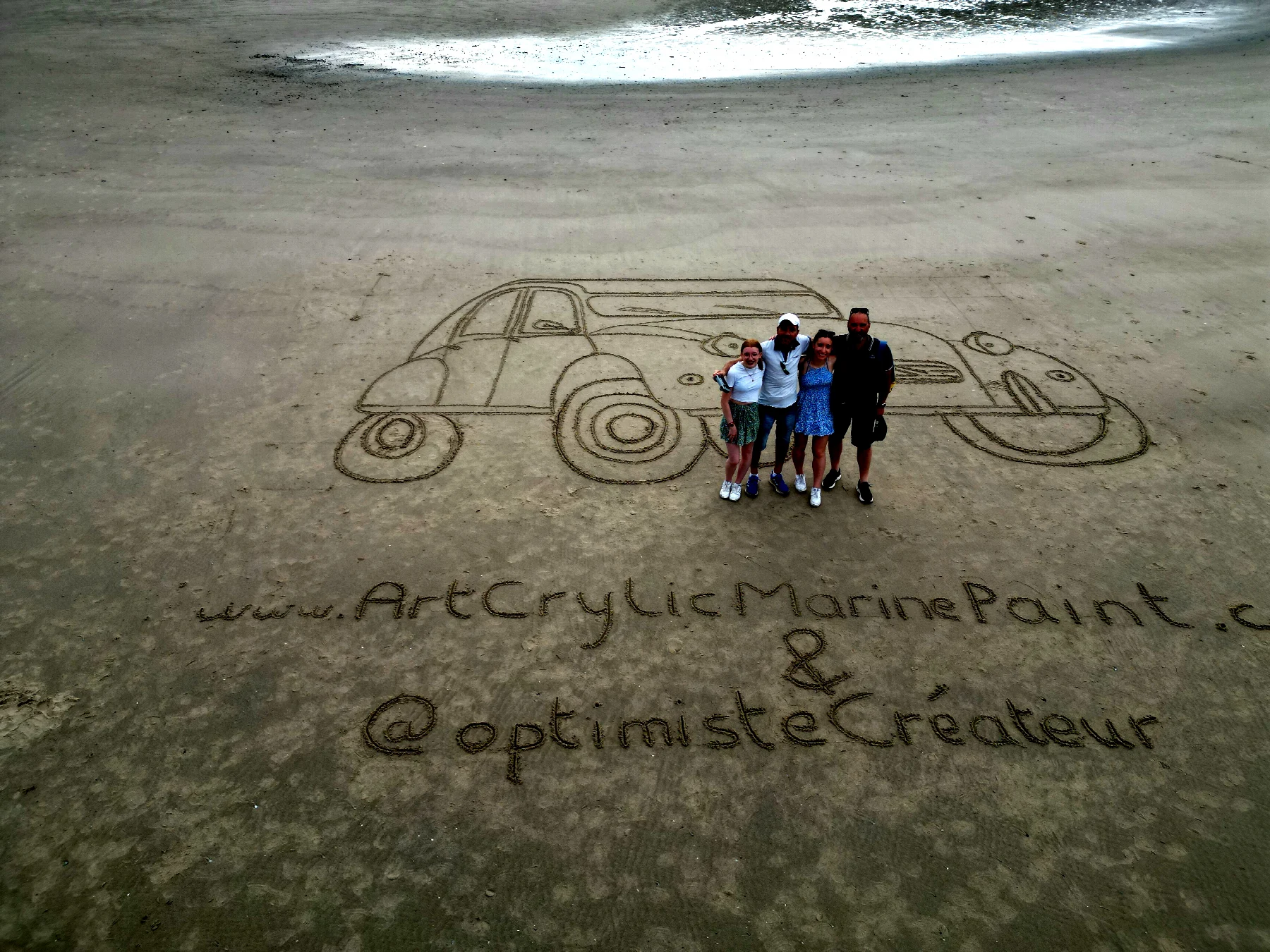 Fresque d’une voiture ancienne tracée dans le sable avec un groupe de personnes devant l’œuvre