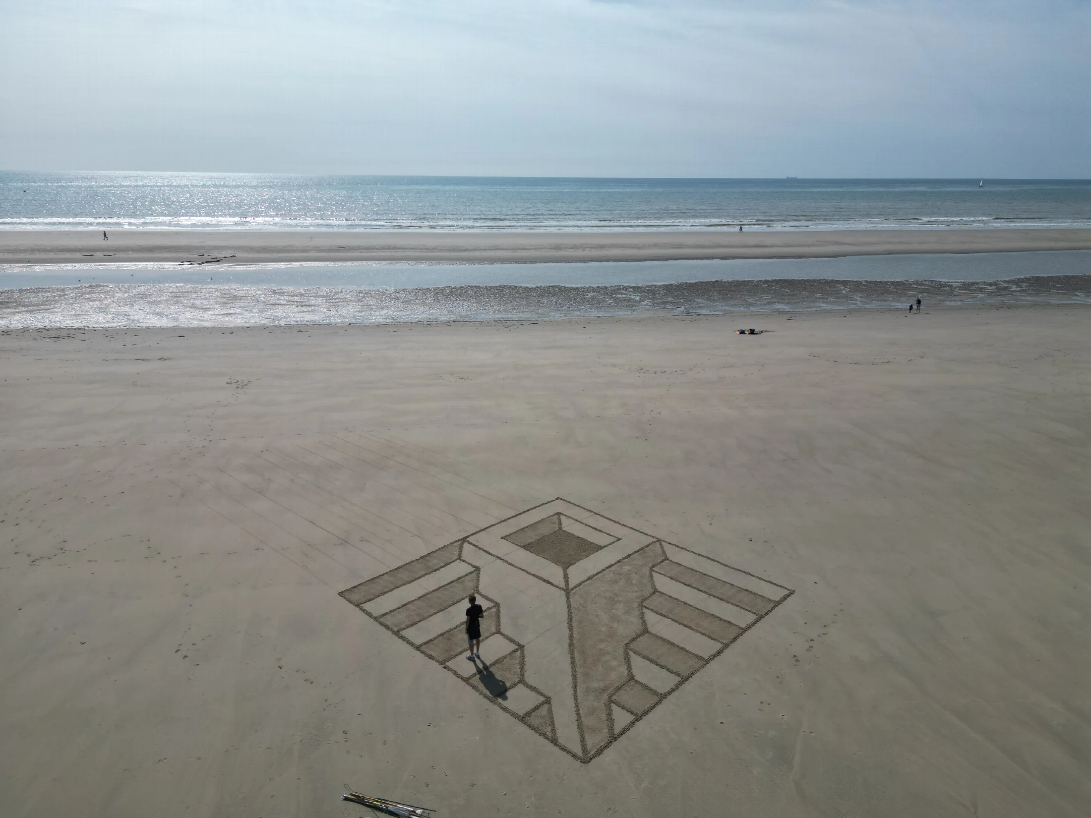 Grande fresque géométrique en perspective tracée dans le sable avec une personne face à la mer
